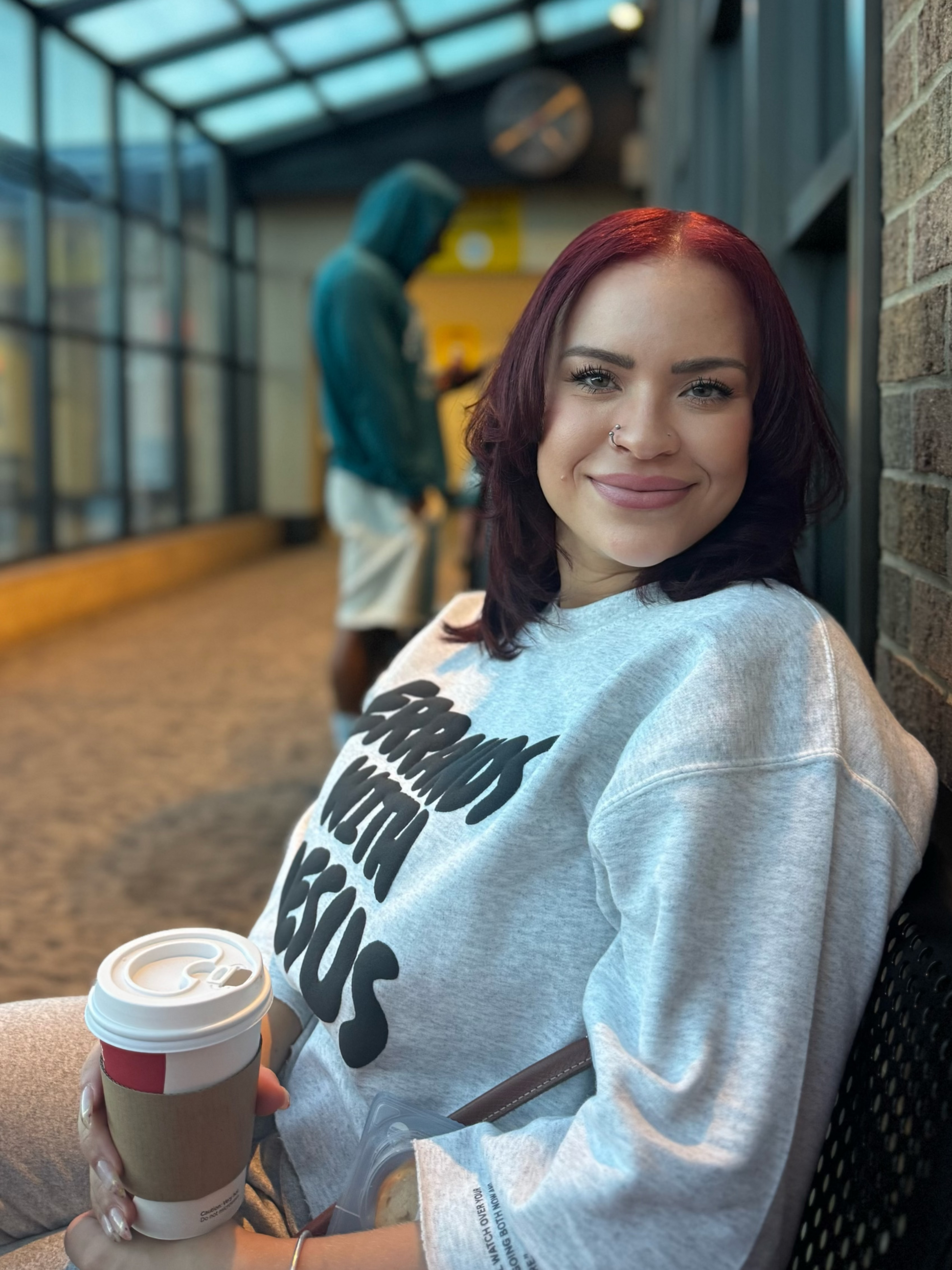 Woman with red hair wearing a sweatshirt with text Errands with Jesus holding a coffee cup, in the airport